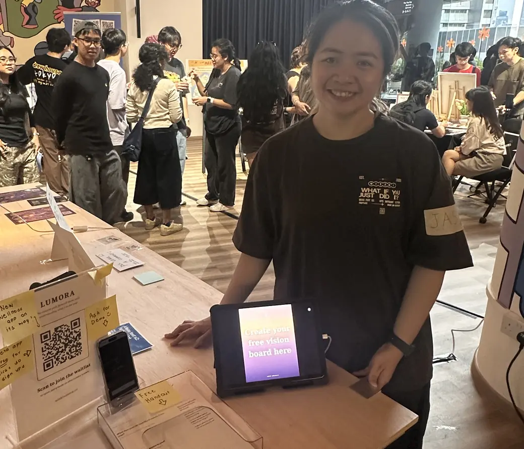 Jas at a Lumora booth, the app running on a tablet beside printed handouts, with a busy room behind.